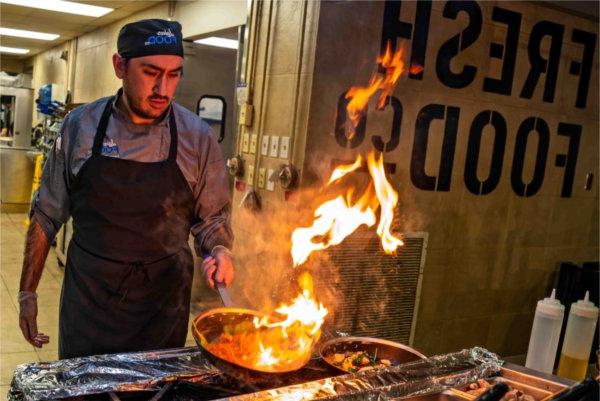 A chef wearing an apron and hat has fire shooting up from a pan as they make food. 