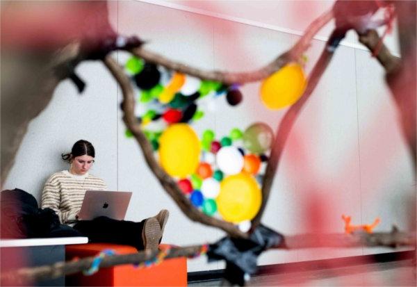  A college student works on their laptop while sitting on an orange bench near colorful artwork. 